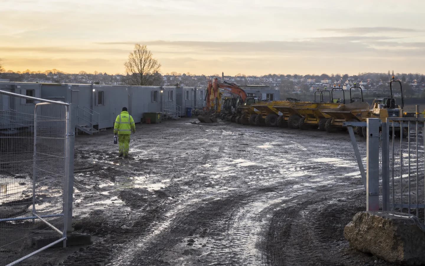 A UK groundworks compound at first light: welfare cabins, parked plant and a muddy haul road